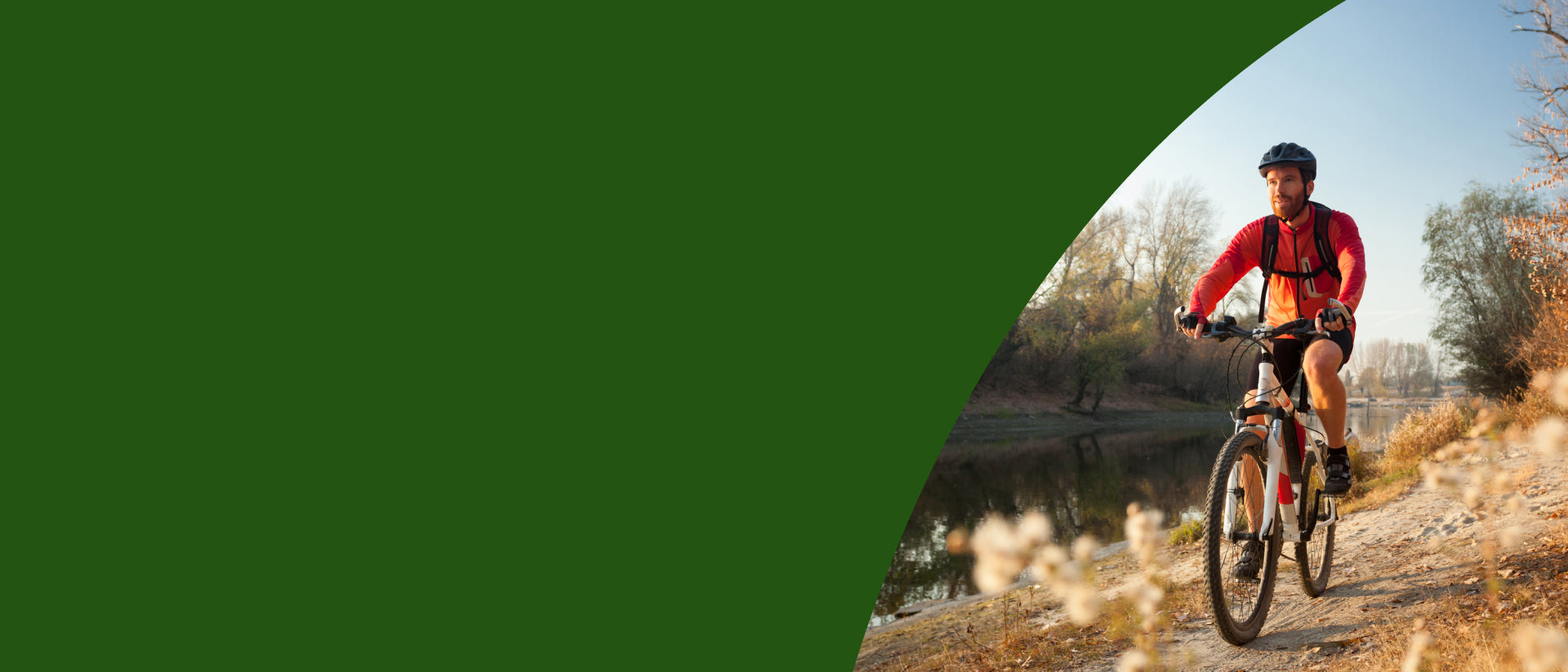 A cyclist wearing a red jacket and black helmet rides a mountain bike on a dirt path next to a river, with a large dark green graphic overlay on the left side of the image - desktop