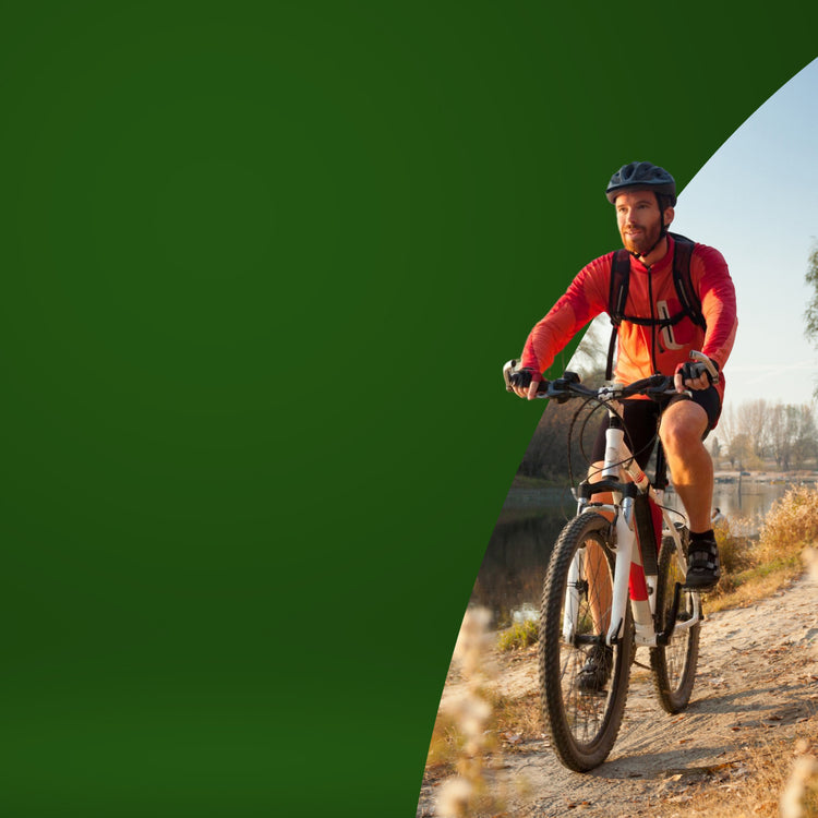 A man wearing a red cycling jersey, black shorts, and a black helmet rides a white and black mountain bike on a dirt path outdoors. The image is split diagonally, with a solid dark green shape on the left and a real outdoor environment on the right - mobile